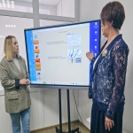 На фото две женщины стоят перед большим интерактивным экраном с презентацией, на котором отображены тестовые задания, включая задания с несколькими вариантами ответов и на установление соответствия. Одна из женщин (в тёмно-синем кружевном жакете) жестикулирует, объясняя материал, вторая (в клетчатом пиджаке) внимательно слушает.