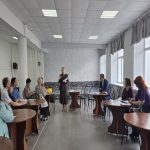 В светлом конференц-зале проходит рабочее совещание: в центре помещения стоит женщина в деловом костюме и с документами в руках, обращаясь к собравшимся. Участники встречи расположились за круглыми деревянными столами по обе стороны зала, внимательно слушая выступающую.