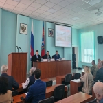В актовом зале проходит официальное мероприятие: за длинным столом на сцене сидят трое мужчин в деловых костюмах, перед ними — таблички с именами, микрофоны и стаканы с водой. В зале присутствуют слушатели, на стене видны флаги России и региона, герб, а на проекционном экране демонстрируется презентация с заголовком «Проект „Мобильный ФАП“».