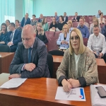 На переднем плане за столом сидят мужчина в сером пиджаке и женщина в клетчатом жакете с шарфом, перед ними лежат документы и ручка; на заднем плане — аудитория с людьми в деловой одежде, сидящими на бордовых креслах в актовом зале с голубыми стенами и вертикальными жалюзи на окнах.