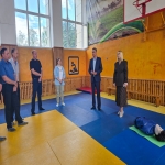 В просторном спортивном зале на разноцветных матах собралась группа из шести человек, внимательно слушающих мужчину в деловом костюме, стоящего в центре. На переднем плане справа лежит манекен для отработки приёмов, а на заднем фоне видны баскетбольное кольцо и тематический баннер с изображением лошадей.