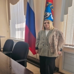 Женщина в клетчатом пиджаке, светлой блузке и с накинутым на плечи платком с узором стоит в официальном помещении. На заднем плане видны флаги России и региона, а также стол с двумя офисными креслами у окна с жалюзи.