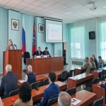 В зале проходит официальное мероприятие: женщина выступает с речью за трибуной, а за длинным столом сидят двое мужчин с микрофонами и табличками с именами. В зале присутствуют слушатели, на стене видны флаги России и региона, а также проекция презентации с заголовком «Реализация проекта „Готов к санитарной обороне“».