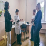 В светлом кабинете четыре женщины ведут беседу: в центре стоит сотрудница в белом медицинском халате и активно жестикулирует, а напротив неё полукругом расположились собеседницы в деловой и повседневной одежде. На заднем плане у стены виден стол с канцелярскими принадлежностями и информационными плакатами, один из которых посвящён профилактике педикулёза.