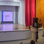 Женщина выступает с докладом, стоя за деревянным подиумом с микрофоном на сцене актового зала. На большом экране позади неё отображается слайд с темой мероприятия — «Реализация проекта „Готов к санитарной обороне“» в Бийском округе.