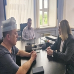 В светлом кабинете за большим столом сидят двое собеседников: мужчина в серой футболке и медицинской шапочке и женщина в деловом пиджаке в полоску, они внимательно смотрят друг на друга, сложив руки. На заднем плане в кресле расположился ещё один мужчина в светлой рубашке, наблюдающий за диалогом, а за окнами видна зелёная листва.