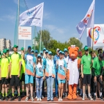 На спортивной площадке собралась большая группа людей в яркой спортивной форме, позирующих для общего фото; в центре композиции выделяется ростовая кукла — медведь в белом медицинском халате и шапочке с красным крестом.  На заднем плане видны флаги и табличка с надписью «КГБПОУ „Алтайский институт развития медицинского образования“», что указывает на принадлежность участников к этому учебному заведению и, вероятно, на проведение корпоративного спортивного мероприятия.
