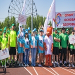 На спортивной арене собралась большая группа участников в яркой спортивной форме, которые позируют для общего фото; среди них выделяется персонаж в костюме плюшевого медведя в медицинской шапочке.  На заднем плане видны баннеры с лозунгом «Спорт — основа здоровья» и логотипами медицинских учреждений, а вдалеке возвышается конструкция колеса обозрения на фоне ясного неба.