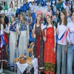 На трибунах спортивного стадиона группа молодых девушек в ярких традиционных русских костюмах позирует с маленькими флагами России, стоя рядом с небольшим столиком, накрытым угощениями — пирогами, самоваром и связками баранок.  На заднем плане видны другие участники праздника с плакатами патриотической тематики и болельщики в повседневной одежде, создающие атмосферу массового торжественного мероприятия.