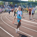 На переднем плане спортсменка в бирюзовой форме с номером 9 стремительно преодолевает дистанцию по легкоатлетической дорожке.  На заднем плане видны другие участники соревнований и многочисленная группа зрителей, расположившихся на трибунах и вдоль беговой зоны.