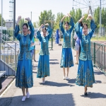 На фотографии четыре девушки исполняют национальный танец на улице, одетые в яркие синие костюмы с золотой вышивкой.  На заднем плане видны элементы городской инфраструктуры — колесо обозрения, спортивная площадка и зелёные деревья, что создаёт атмосферу массового культурного или праздничного мероприятия.