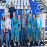 На групповом фото на фоне пустых сине-белых трибун стадиона позирует разновозрастная компания людей.  В центре группы — мужчины в повседневной одежде, а по бокам от них выделяются девушки в традиционных бирюзовых костюмах с богатой золотой вышивкой.