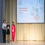 На сцене проходит торжественное собрание, посвящённое Дню медицинского работника, — об этом сообщает большой экран с надписью «Медицина начинается здесь» и логотипом Алтайского института развития медицинского образования.  В центре внимания — женщина в ярко-розовом костюме, выступающая с микрофоном; по бокам от неё стоят двое молодых людей с листами бумаги и микрофонами, готовые к выступлению.
