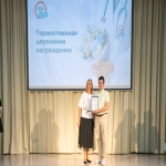 На сцене проходит торжественная церемония награждения, организованная Алтайским институтом развития медицинского образования — это видно по надписи и логотипу на большом экране позади выступающих.  В центре кадра женщина в белой блузке и чёрной юбке вручает мужчине в светлой рубашке и брюках сертификат в рамке.