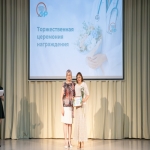 На сцене проходит торжественная церемония награждения, организованная Алтайским институтом развития медицинского образования — это подтверждает надпись и логотип на большом экране позади выступающих.  Две женщины позируют для фото: одна вручает другой сертификат в рамке, а награждённая держит в руках букет белых цветов.