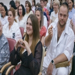 На переднем плане молодая женщина в чёрном платье в красный горошек улыбается и складывает руки в форме сердца, сидя в зале среди коллег.  Рядом с ней мужчина в белом медицинском халате смотрит в сторону и делает жест пальцем, на его запястье видны смарт-часы.