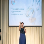 На сцене проходит торжественная церемония награждения, организованная Алтайским институтом развития медицинского образования — это подтверждает надпись и логотип на большом экране позади выступающей.  В центре внимания женщина в тёмно-синем наряде, которая держит в руке микрофон и эмоционально жестикулирует, обращаясь к аудитории.