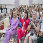 В зале проходит торжественное мероприятие: зрители, преимущественно женщины, активно аплодируют и снимают происходящее на смартфоны.  На переднем плане выделяются три участницы в ярких костюмах — сиреневом, розовом и с пестрым принтом — они улыбаются и с энтузиазмом поддерживают выступающих.