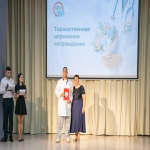 На сцене Алтайского института развития медицинского образования проходит торжественная церемония награждения, о чём свидетельствует крупный экран с тематическим оформлением на заднем плане.  В центре внимания — мужчина в белом медицинском халате, держащий в руках красную папку с наградой, рядом с ним стоит женщина в тёмно-синем платье.