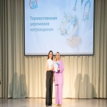 На сцене Алтайского института развития медицинского образования проходит торжественная церемония награждения, о чём свидетельствует крупный экран с тематическим оформлением на заднем плане.  Две женщины — одна в белом топе и чёрных брюках, другая в светло-лиловом костюме — стоят рядом, держа в руках сертификат, и тепло улыбаются в камеру.