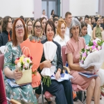 В зале собрались женщины — участницы торжественного мероприятия: они держат в руках букеты цветов и папки с документами, на лицах — спокойные улыбки.