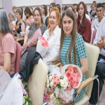 В зале собрались женщины — участницы торжественного мероприятия: они держат в руках букеты цветов и папки с документами, на лицах — спокойные улыбки.