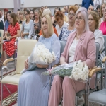 В зале собрались женщины — участницы торжественного мероприятия: они держат в руках букеты цветов и папки с документами, на лицах — спокойные улыбки.