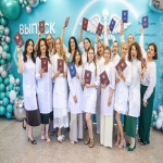 Группа выпускниц медицинского учреждения в белых халатах радостно позирует с дипломами на фоне праздничной фотозоны с надписью «ВЫПУСК» и символикой медицинской профессии.