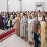 В просторном светлом зале собралась большая группа людей в деловой и полуформальной одежде — они выстроились в ряды, вероятно, для общего фото или участия в торжественном мероприятии.