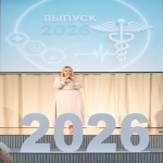 На сцене женщина в светло‑бежевом платье с микрофоном выступает на торжественном мероприятии, посвящённом выпуску 2026 года, — на фоне экрана с медицинской символикой и крупными цифрами «2026» на переднем плане.