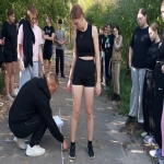 На изображении девушка в чёрном платье стоит на улице, рядом с ней человек на корточках. Вокруг люди, деревья и кусты.