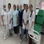 На изображении группа людей в медицинской одежде стоит в помещении. Справа зелёное кресло-каталка. Люди улыбаются и позируют для фото.