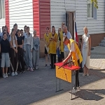 На изображении группа людей, стоящих на улице возле здания. Один человек перерезает ленточку, символизируя открытие или праздник.