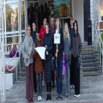 На изображении группа людей, стоящих на улице. Большинство из них одеты в тёмную одежду. На заднем плане здание