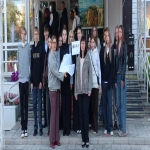 На изображении группа людей, стоящих на улице. Большинство из них одеты в тёмную одежду. На заднем плане здание
