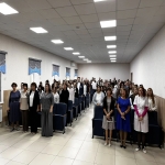 На изображении большое помещение с потолочными светильниками. В комнате много людей, стоящих рядами. Пол светлый, стены светлые.