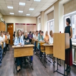 В помещении с ярким освещением группа людей сидит за партами. Одна женщина стоит у доски и что-то объясняет.