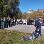 На изображении видна группа людей, стоящих на улице в парке. На переднем плане человек ухаживает за лежащим на траве. Погода ясная, небо голубое.