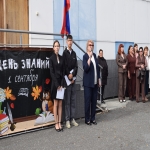 На изображении группа людей на улице, некоторые в костюмах, рядом флаг, на заднем плане надпись 