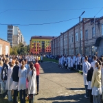 На изображении видна улица с людьми в белых халатах. На заднем плане видны здания и голубое небо.