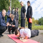 На изображении группа людей на улице. Один человек лежит на коврике, другие стоят вокруг. Вокруг зелёная растительность и деревья.