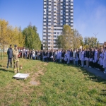 На изображении видно зелёную лужайку, группу людей, стоящих в ряд, и высокие здания на заднем плане. Погода солнечная, небо ясное.