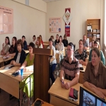 В помещении сидят люди за партами, слушают выступающего. На стенах висят плакаты. Освещение естественное, дневное.