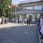 На изображении видна площадь перед зданием, справа стоят люди в белых халатах. В центре площади несколько человек. Слева деревья и флагштоки.