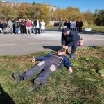 На изображении человек лежит на траве, рядом стоят другие люди. На заднем плане видны деревья и группа людей. Погода солнечная.