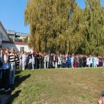 На изображении видна группа людей, стоящих на траве возле деревьев. На заднем плане видны здания и голубое небо.