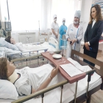 На изображении кабинет - больничная палата с несколькими кроватями, на которых лежат манекены, накрытые белыми простынями. Рядом стоят люди в медицинской одежде и костюме.