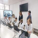 В комнате за столами сидят люди с ноутбуками, рядом стоят преподаватель. На стене висит телевизор.
