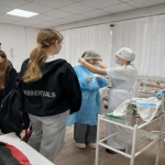 На изображении несколько человек, стоящих в помещении. Видны манекены в одежде, стол с предметами и белая занавеска на заднем плане.