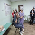 На изображении человек сидит на стуле, рядом медсестра в халате. В комнате также находятся несколько людей, наблюдающих за происходящим.